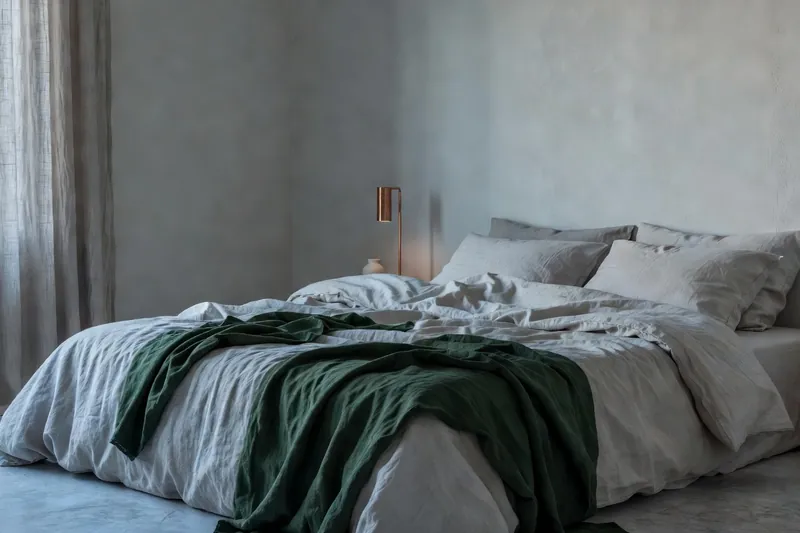 Chambre au petit matin, draps de lin clair défaits et rideau filtrant une lumière bleutée.