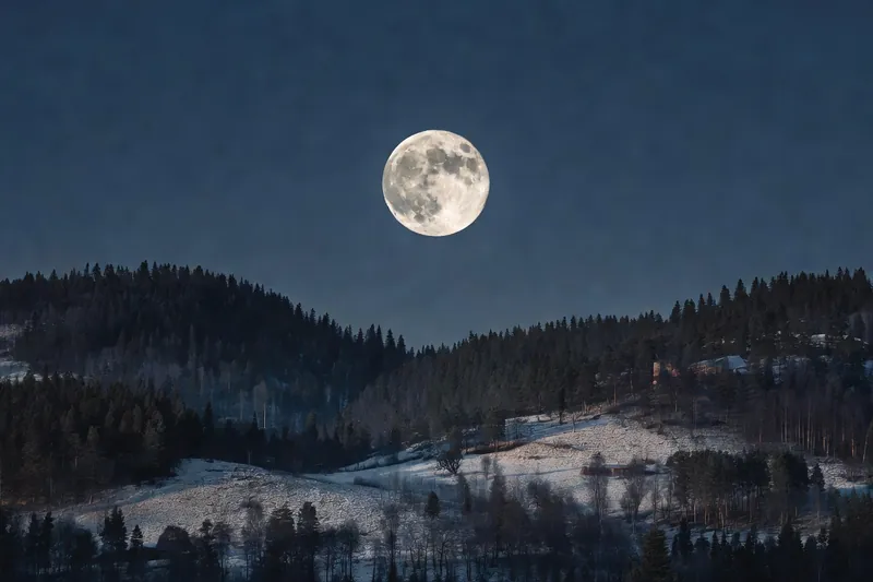 Pleine Lune détaillée au-dessus de collines boisées sous un ciel nocturne dégagé.