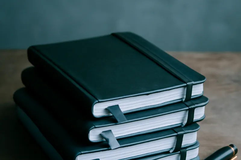 Carnets de terrain empilés, marque-pages colorés et stylo sur un bureau en bois usé.